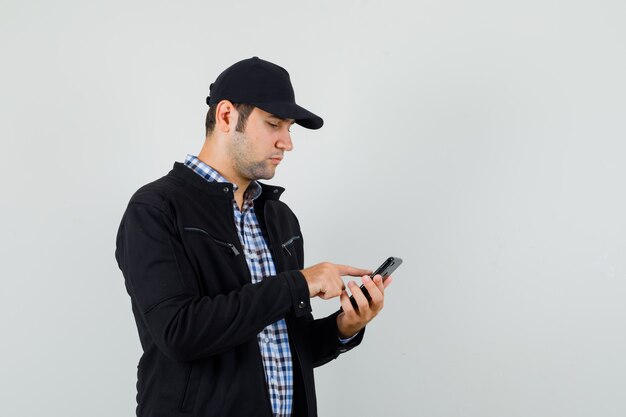 Молодой человек с помощью мобильного телефона в рубашке, куртке, кепке и выглядит занятым, вид спереди.