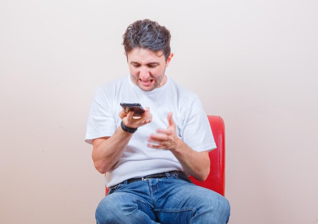 Young man using mobile phone while sitting on chair in t-shirt, jeans and looking angry