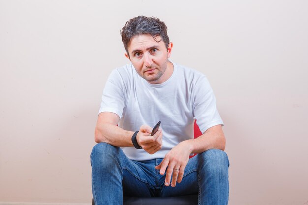 T- 셔츠와 청바지에 의자에 앉아있는 동안 원격 컨트롤러를 사용하는 젊은 남자