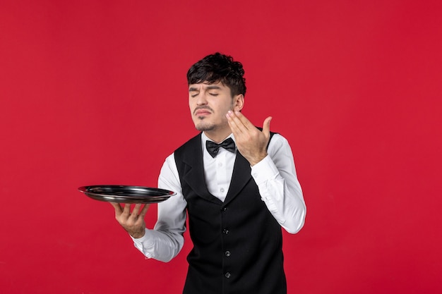 young man waiter in a uniform tying butterfly on neck holding tray smelling on isolated red background