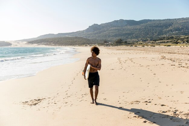 Young man walking along seashore carrying surfboard 