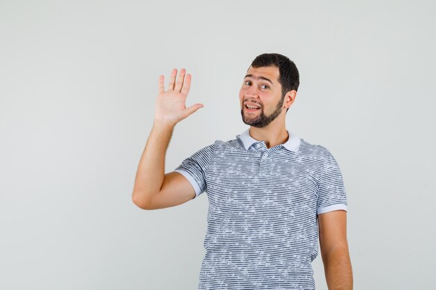 Young man waving hand for greeting in t-shirt and looking merry , front view.