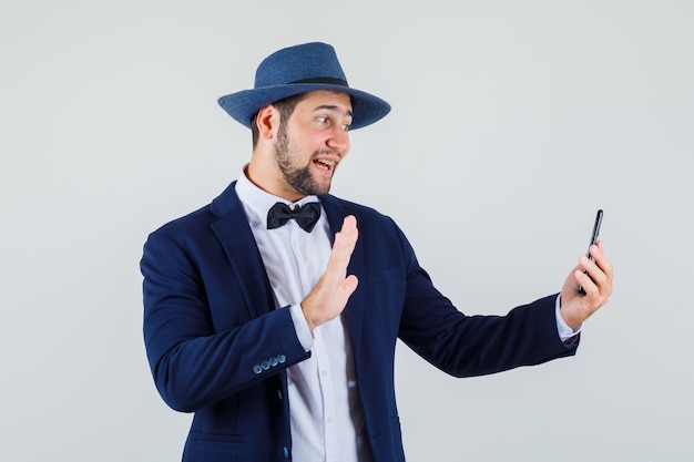 Young man waving hand while making video call in suit, hat and looking optimistic , front view.