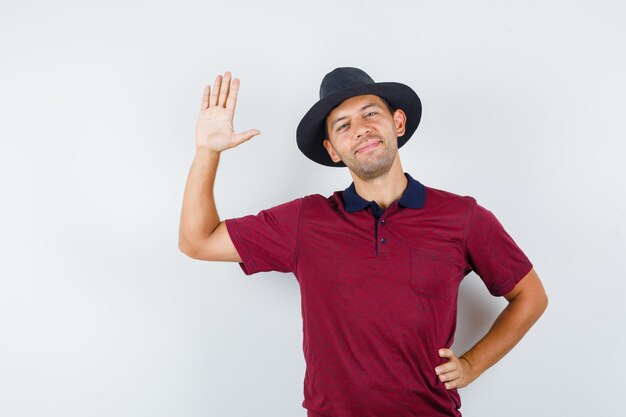 Молодой человек машет ладонью, чтобы поздороваться в футболке, шляпе и выглядит радостным, вид спереди.