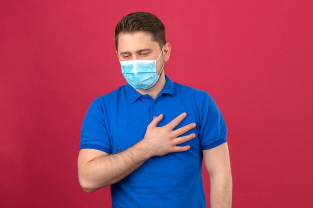 Young man wearing blue polo shirt in medical protective mask holding his hand on chest lungs feeling bad standing over isolated pink wall