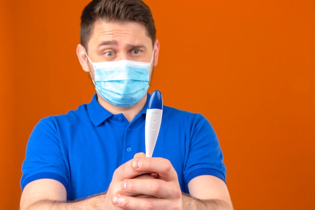 緊張と分離のオレンジ色の壁を心配している手でデジタル温度計を見て医療用防護マスクで青いポロシャツを着ている若い男