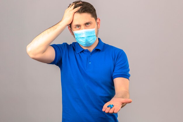 Молодой человек в синей рубашке поло в медицинской защитной маске выглядит нездоровым и больным, стоя с рукой на голове и страдает от головной боли, держа пилинги в открытой руке над изолированной оранжевой стеной