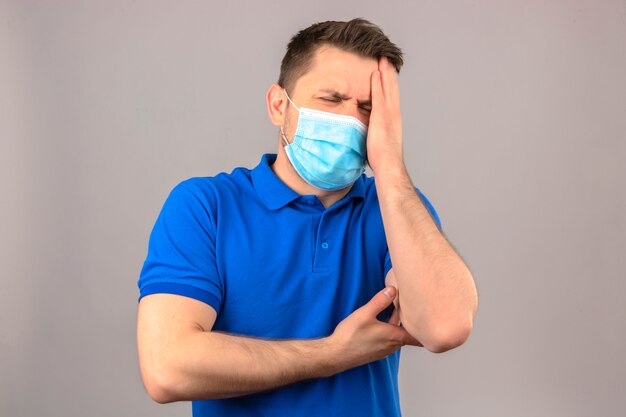 Young man wearing blue polo shirt in medical protective mask looking unwell and sick standing with hand on head suffering from headache over isolated white wall