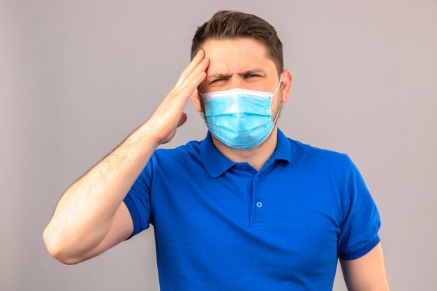 Молодой человек в синей рубашке поло в медицинской защитной маске выглядит нездоровым и больным, стоя с рукой на голове и страдает от головной боли над изолированной белой стеной