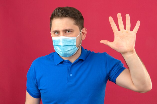 Young man wearing blue polo shirt in medical protective mask making greeting gesture with open hand waving standing over isolated pink wall