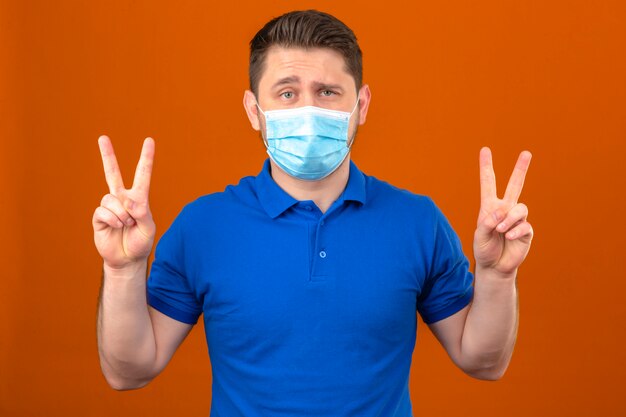 Молодой человек в синей рубашке поло в медицинской защитной маске показывает знаки победы с пальцами, стоящими над изолированной оранжевой стеной
