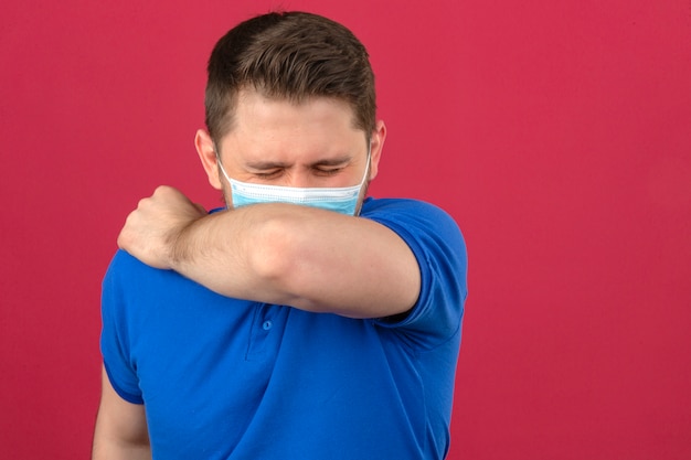 分離されたピンクの壁にcovid-19coronavirusが広がるのを防ぐために腕または肘に咳をするくしゃみをする医療用防護マスクで青いポロシャツを着ている若い男