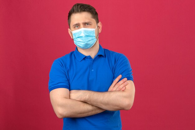 Молодой человек в синей рубашке поло в медицинской защитной маске стоит со скрещенными руками и уверенно смотрит на изолированную розовую стену