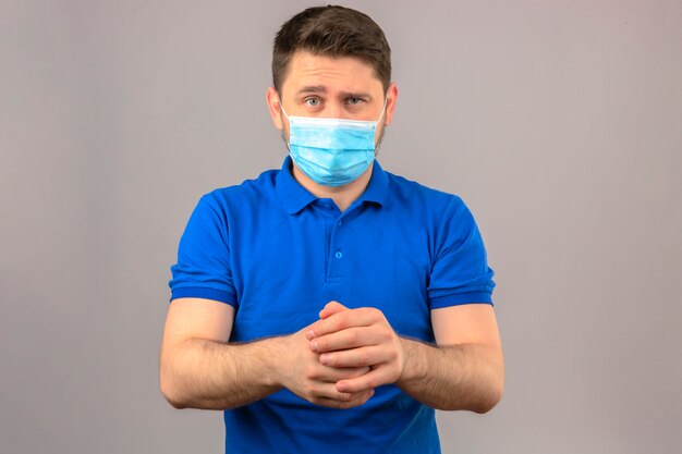 Young man wearing blue polo shirt in medical protective mask with skeptic expression holding hands together standing over isolated white wall