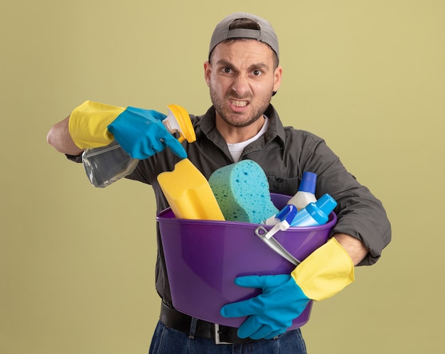 Young man wearing casual clothes and cap in rubber gloves holding bucket with cleaning tools and cleaning spray lwith angry face being displeased ooking  standing over green wall