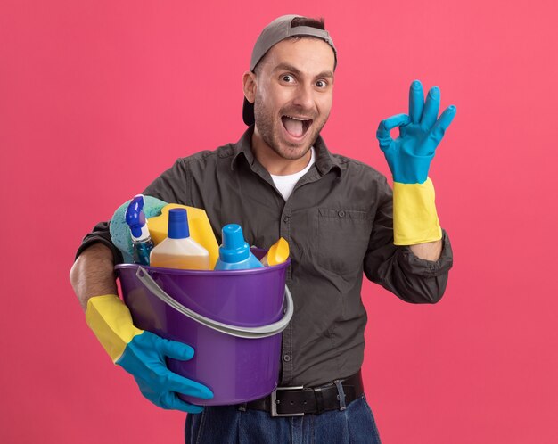 Young man wearing casual clothes and cap in rubber gloves holding bucket with cleaning tools looking  smiling cheerfully showing ok sign standing over pink wall