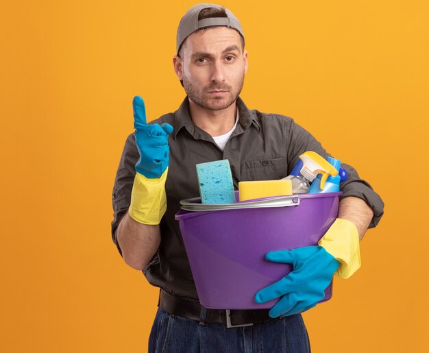 Young man wearing casual clothes and cap in rubber gloves holding bucket with cleaning tools looking  with confident expression showing index finger standing over orange wall