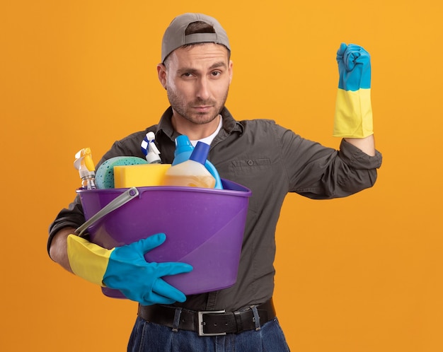 Young man wearing casual clothes and cap in rubber gloves holding bucket with cleaning tools looking  with serious face raising fist standing over orange wall