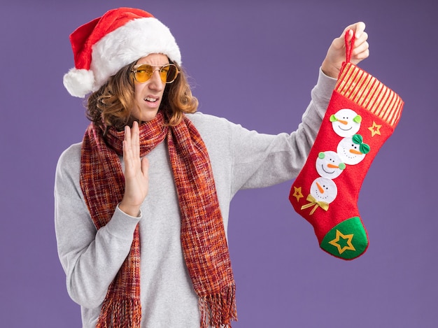 Бесплатное фото Молодой человек в рождественской шапке санта-клауса и желтых очках с теплым шарфом на шее держит рождественский чулок, глядя на него с отвращением на фиолетовом фоне