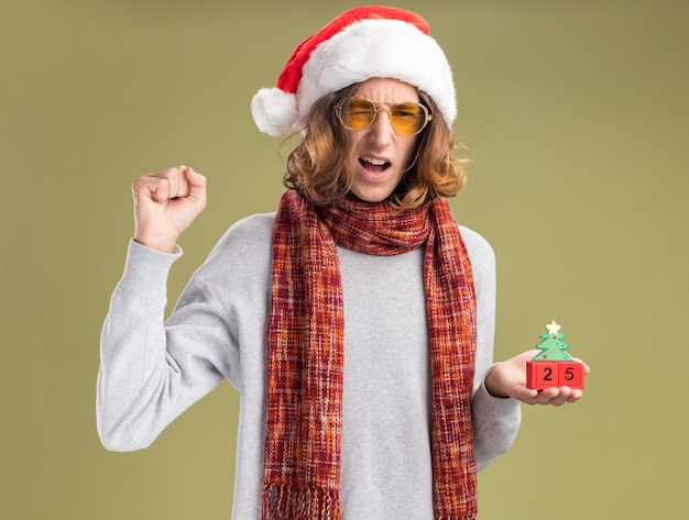 녹색 배경 위에 행복하고 흥분 서 날짜 25 떨림 주먹으로 장난감 큐브를 들고 그의 목에 따뜻한 스카프와 함께 크리스마스 산타 모자와 노란색 안경을 착용하는 젊은 남자