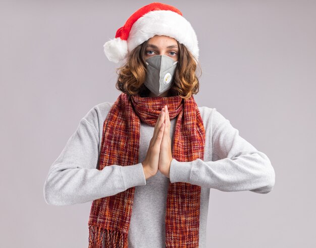 Young man wearing christmas santa hat and facial protective mask  with warm scarf around his neck holding hands together  with serious face standing over white  wall