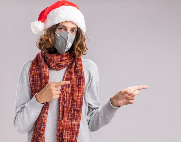 young man wearing christmas santa hat and facial protective mask with warm scarf around his neck looking at camera pointing with index fingers to the side standing over white background