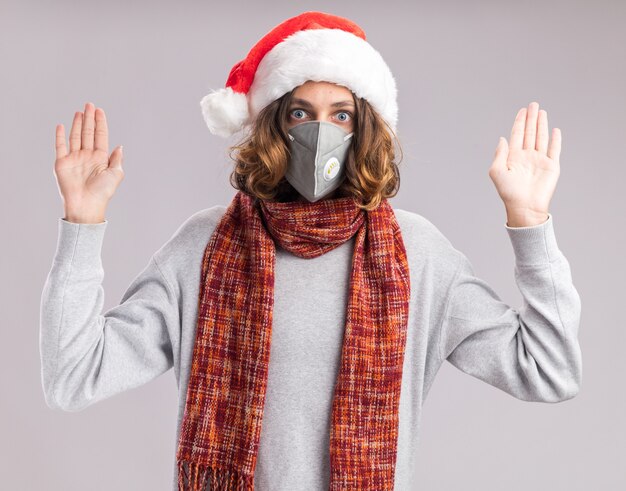 Young man wearing christmas santa hat and facial protective mask  with warm scarf around his neck  worried raising hands in surrender standing over white  wall