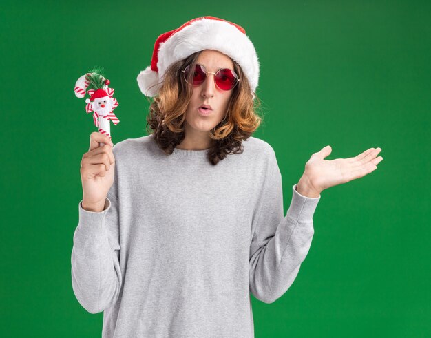 Молодой человек в рождественской шляпе санта-клауса и красных очках держит рождественскую конфету, глядя в камеру, смущенный представлением с рукой, стоящей на зеленом фоне