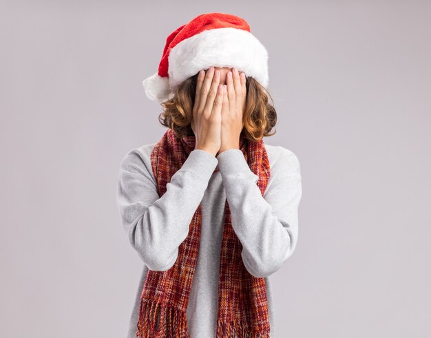 Giovane uomo che indossa il cappello di babbo natale con sciarpa calda intorno al collo che copre il viso con le mani