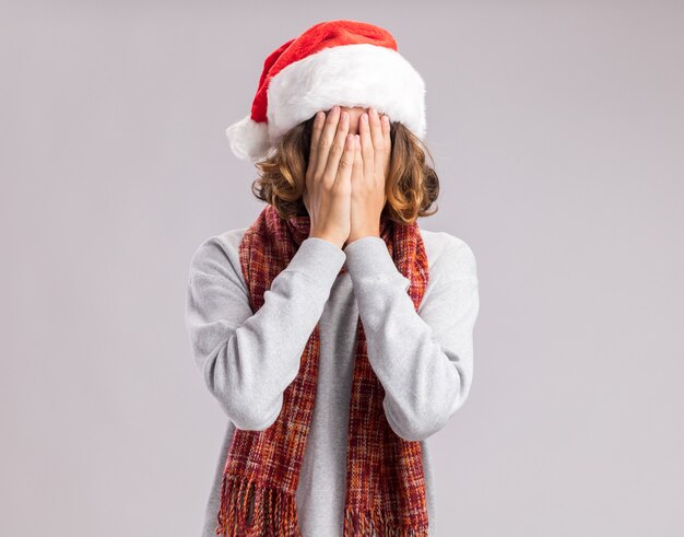 Молодой человек в рождественской шапке санта-клауса с теплым шарфом на шее, закрывая лицо руками