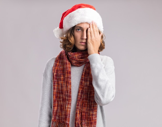 young man wearing christmas santa hat with warm scarf around his neck looking at camera with serious face covering one eye with hand standing over white background