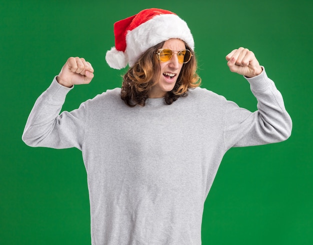 Young man wearing christmas santa hat and yellow glasses clenching fists happy and excited standing over green background