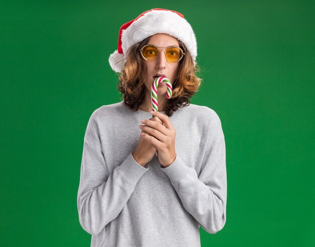 緑の背景の上に立って驚いたカメラを見てキャンディケインを保持しているクリスマスのサンタ帽子と黄色いメガネを身に着けている若い男