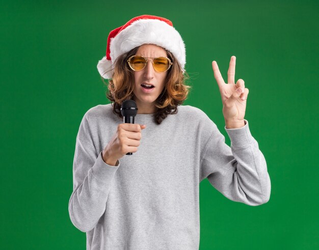 Молодой человек в рождественской шляпе санта-клауса и желтых очках держит микрофон, улыбаясь, показывая v-знак, стоящий над зеленой стеной