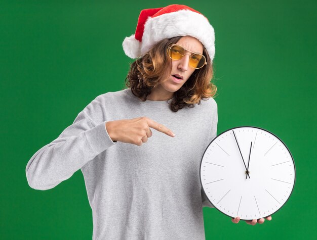 クリスマスのサンタの帽子と黄色いメガネを身に着けている若い男は、緑の壁の上に立って混乱しているように見える人差し指で指している壁時計を保持しています