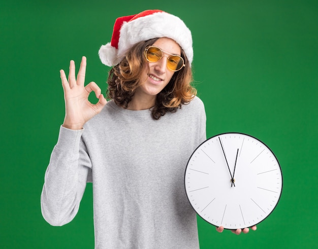 크리스마스 산타 모자와 벽 시계를 들고 노란색 안경을 착용하는 젊은 남자가 녹색 벽 위에 서있는 확인 서명을 보여주는 미소