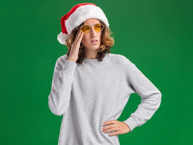 Young man wearing christmas santa hat and yellow glasses looking aside puzzled standing over green background