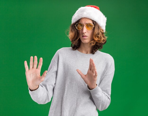 Young man wearing christmas santa hat and yellow glasses looking at camera worried holding hands out standing over green background