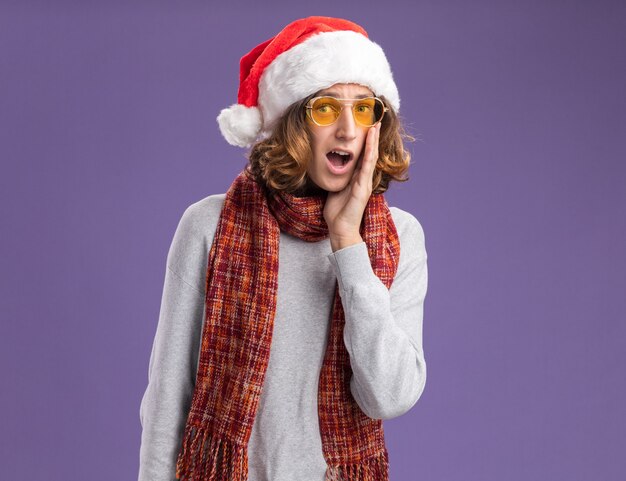 Young man wearing christmas santa hat and yellow glasses with warm scarf around his neck  amazed and surprsied standing over purple  wall