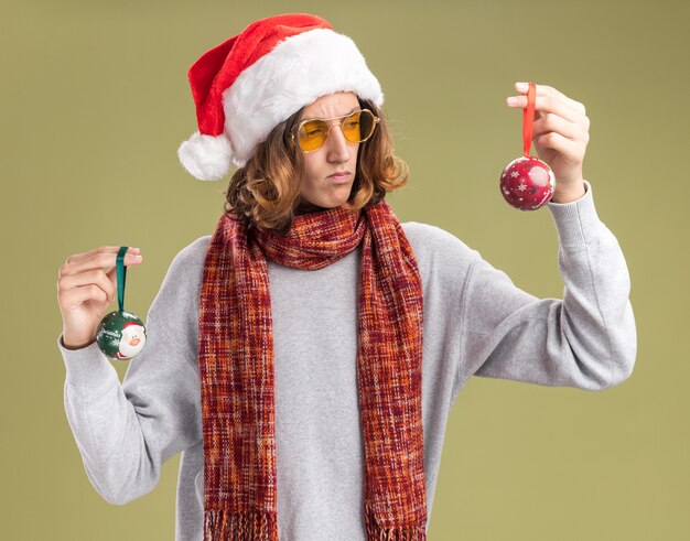 young man wearing christmas santa hat and yellow glasses with warm scarf around his neck holding christmas balls looking confused trying to make choice standing over green background