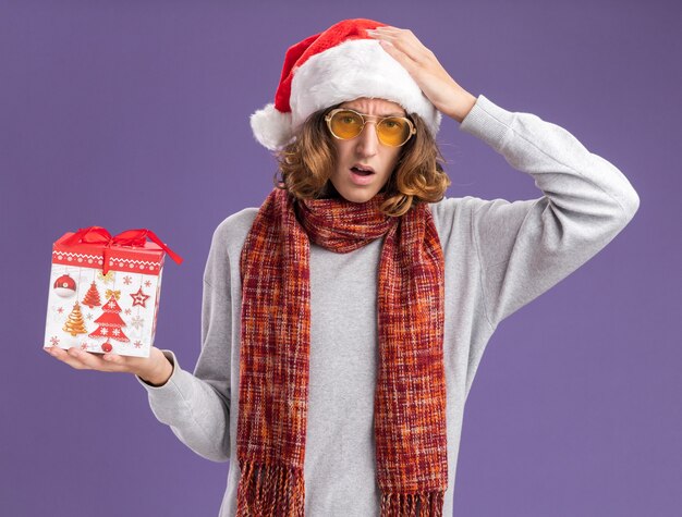 Young man wearing christmas santa hat and yellow glasses with warm scarf around his neck holding christmas gift looking confused with hand over head  standing over purple  background