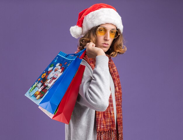 Young man wearing christmas santa hat and yellow glasses with warm scarf around his neck holding christmas paper bags with gifts  with serious face standing over purple  wall