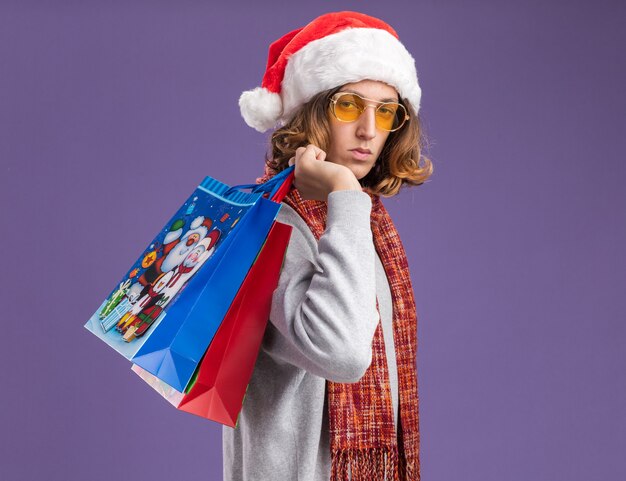Молодой человек в рождественской шляпе санта-клауса и желтых очках с теплым шарфом на шее держит рождественские бумажные пакеты с подарками с серьезным лицом, стоящим над фиолетовой стеной