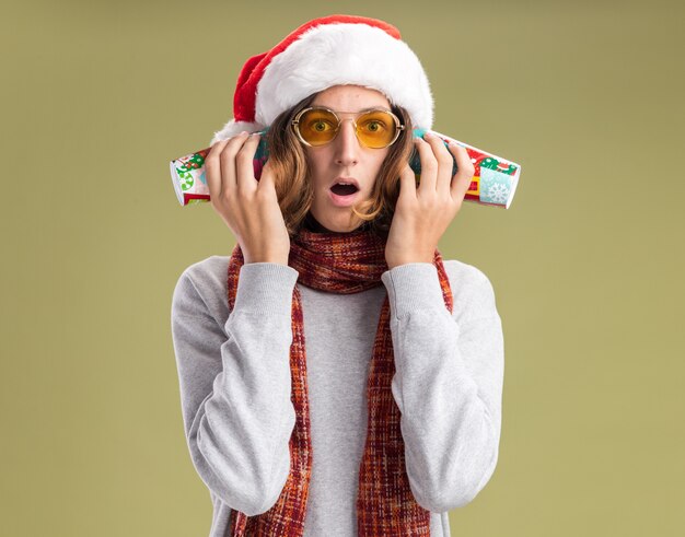 クリスマスのサンタの帽子と黄色いメガネを身に着けている若い男が首に暖かいスカーフを持って、緑の壁の上に立って驚いたように見える彼の耳にカラフルな紙コップを持っています