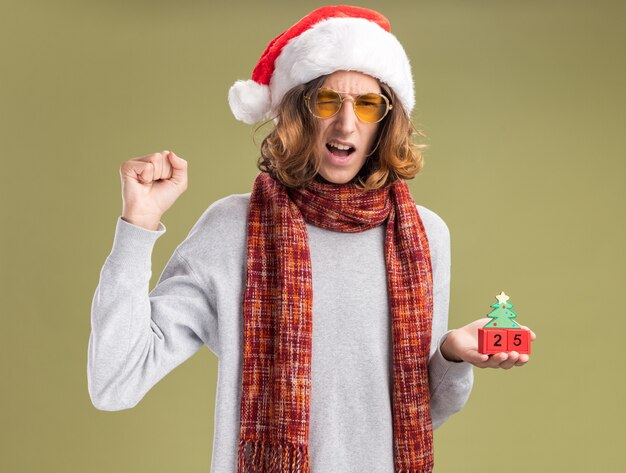 young man wearing christmas santa hat and yellow glasses with warm scarf around his neck holding toy cubes with date twenty five clenching fist happy and excited standing over green background