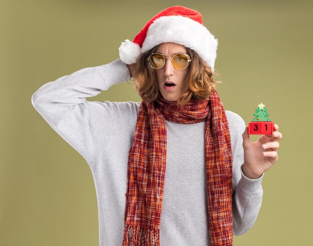 クリスマスのサンタの帽子と黄色いメガネを身に着けている若い男が首に暖かいスカーフを持っておもちゃの立方体を持って、緑の背景の上に立って心配して脇を見ている