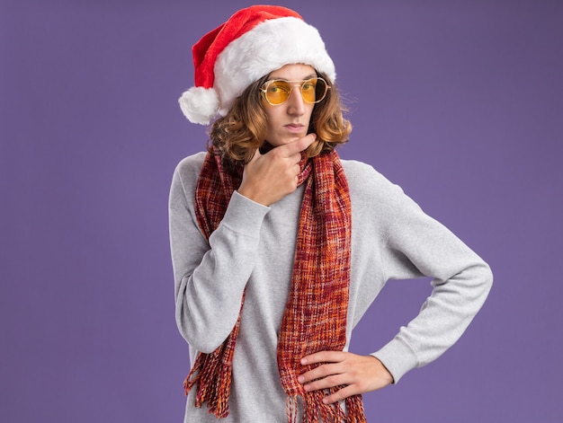 보라색 배경 위에 서있는 회의적인 표정으로 카메라를보고 그의 목에 따뜻한 스카프와 함께 크리스마스 산타 모자와 노란색 안경을 착용하는 젊은 남자