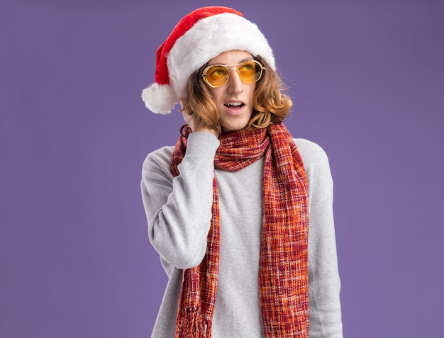 Young man wearing christmas santa hat and yellow glasses with warm scarf around his neck looking up with pensive expression thinking positive standing over purple  wall