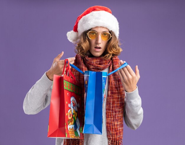 Young man wearing christmas santa hat and yellow glasses with warm scarf around his neck opening  christmas paper bags with gifts  surprised standing over purple  wall