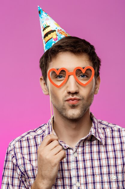 Young man wearing fake glasses over purple wall. Birthday party.